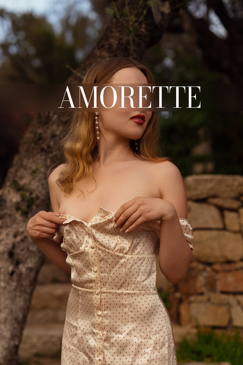 Evoto elite escort model celine stockholm from the side while standing in a nude dress, her blonde hair open and wavy, red lipstick on her lips