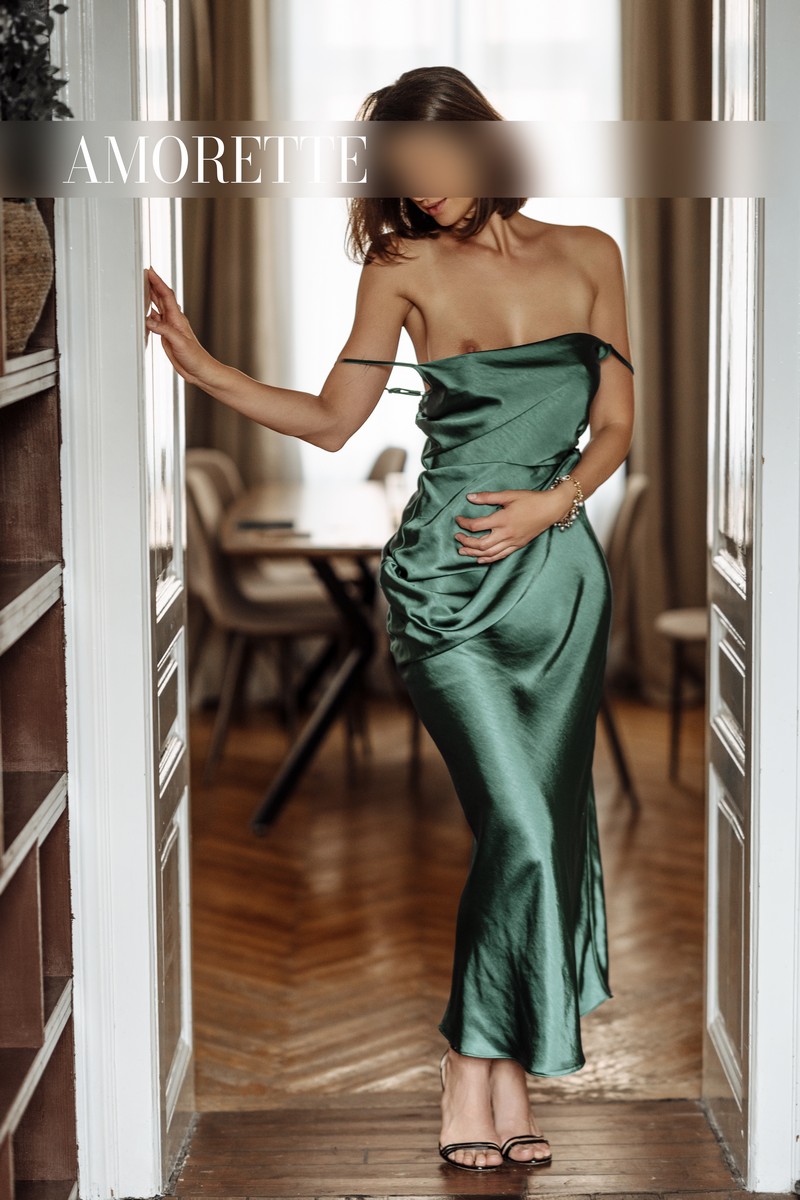 gabrielle elite escort model from vienna shows her from the front while standing next to the door wearing a long green dress, pulling her dress down and showing her natural breasts completely naked, tanned skin, naked breasts, natural breasts, nipples, elegant dress, high heels, short brown hair, high class escort service, austria, vienna, model, lady, luxury, traveling.