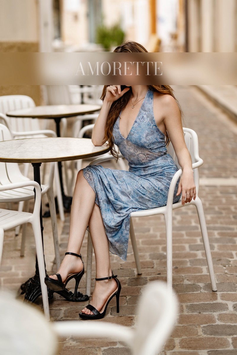 Ellie – Elite Escort Model from Florida Elite escort model Ellie from Florida sits gracefully at a sunlit outdoor terrace, the paved walkway and soft architectural tones providing a sophisticated backdrop. She wears a chic light-blue patterned halter dress that elegantly frames her polished silhouette, one leg crossed over the other in a poised, confident posture. Her long hair cascades softly over her shoulders, while one hand lightly touches her face in a gentle, refined gesture. Black high-heeled sandals with delicate ankle straps complete her ensemble, emphasizing her elegant presence. Soft natural light illuminates her figure and the textures of her outfit, enhancing the luxurious, relaxed, and international atmosphere she exudes. High-class escort model, luxury, traveling, USA, America, international, elegant lingerie, lingerie, models.