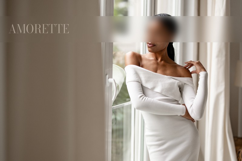 Naomi – Elite Escort Model Stuttgart & Berlin High‑class escort Naomi from Stuttgart & Berlin, with sleek dark hair and a graceful presence, stands elegantly next to a large window in a bright, minimalist interior. She wears a form-fitting white dress that accentuates her refined silhouette, one hand resting softly on her shoulder in a gentle, alluring gesture. Her gaze is directed thoughtfully out the window, adding a serene and sophisticated mood to her poised stance. Soft natural light filters through the glass, highlighting the smooth curves of her figure and casting delicate, flattering shadows. The pristine, elegant backdrop emphasizes her polished beauty, elegance, and premium international appeal. Luxurious, captivating, travel-ready, premium escort, Germany.